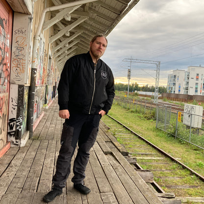Rurik Fightgear-lentäjänpusakka.
Rurik FG "R" -logo rintapielessä sekä riimulogo selässä monivärisesti brodeerattuna. Kuvassa pilottitakki  Mikan päällä junan laiturilla. ptcombet.com